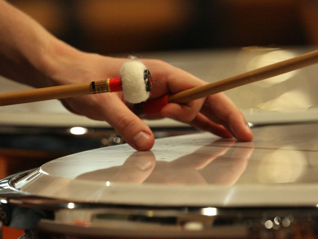 A close-up of a timpanist's hand, muting the skin whilst holding a mallet, the other mallet poised to strike again. Light reflect off the instrument.