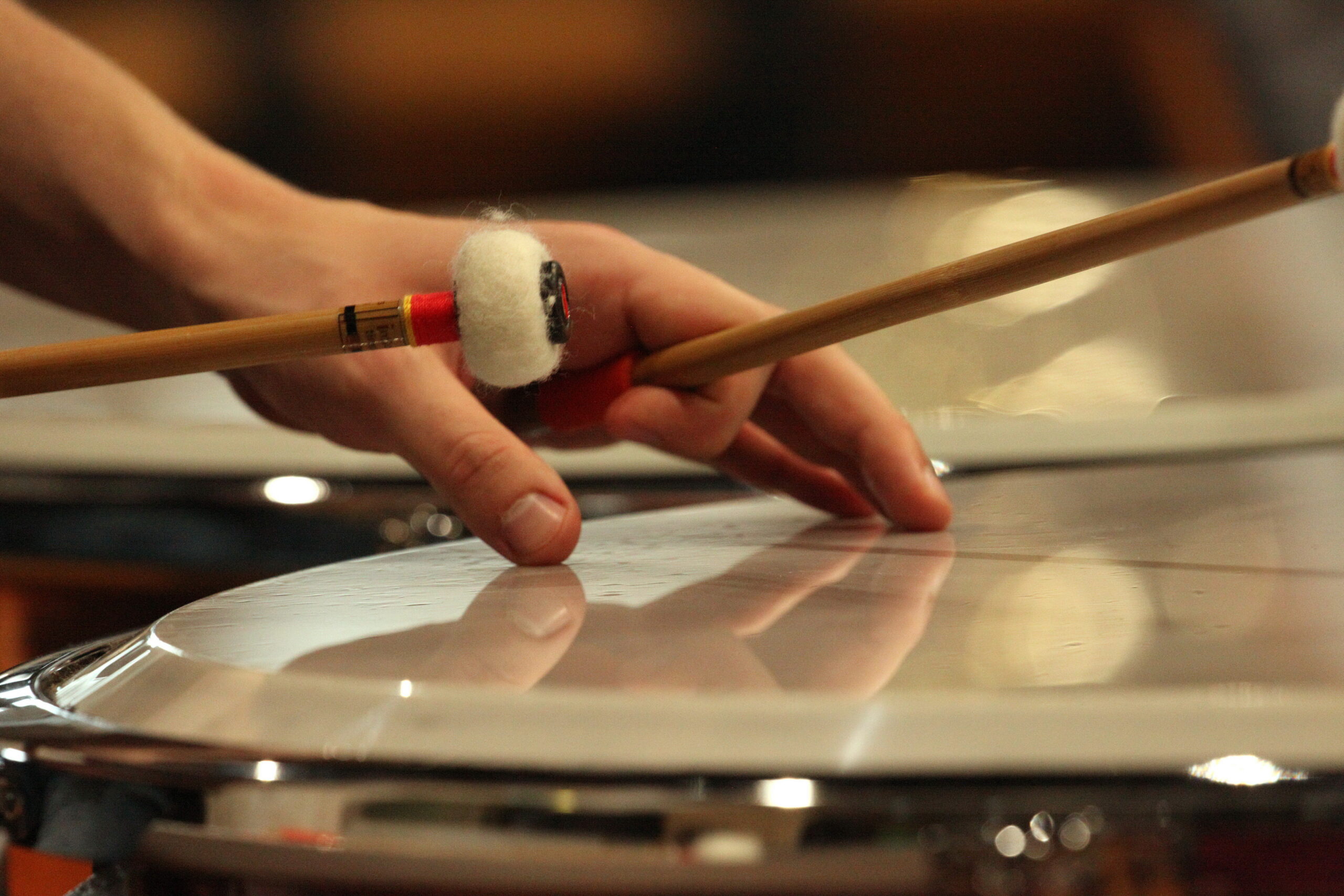 A close-up of a timpanist's hand, muting the skin whilst holding a mallet, the other mallet poised to strike again. Light reflect off the instrument.