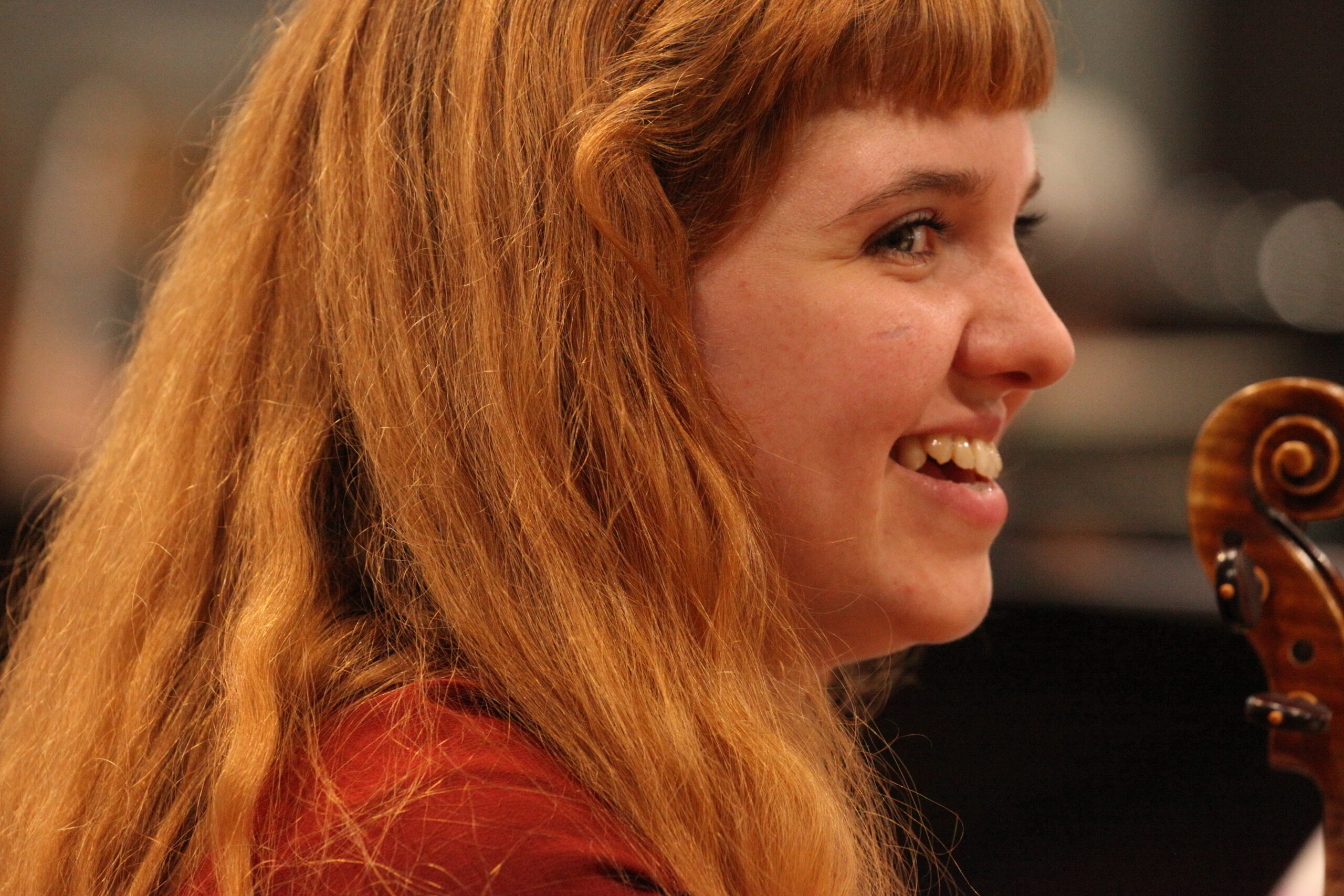 A red-haired violin student at rehearsal, laughing during a break in play. The scroll of her instrument is visible in the corner.