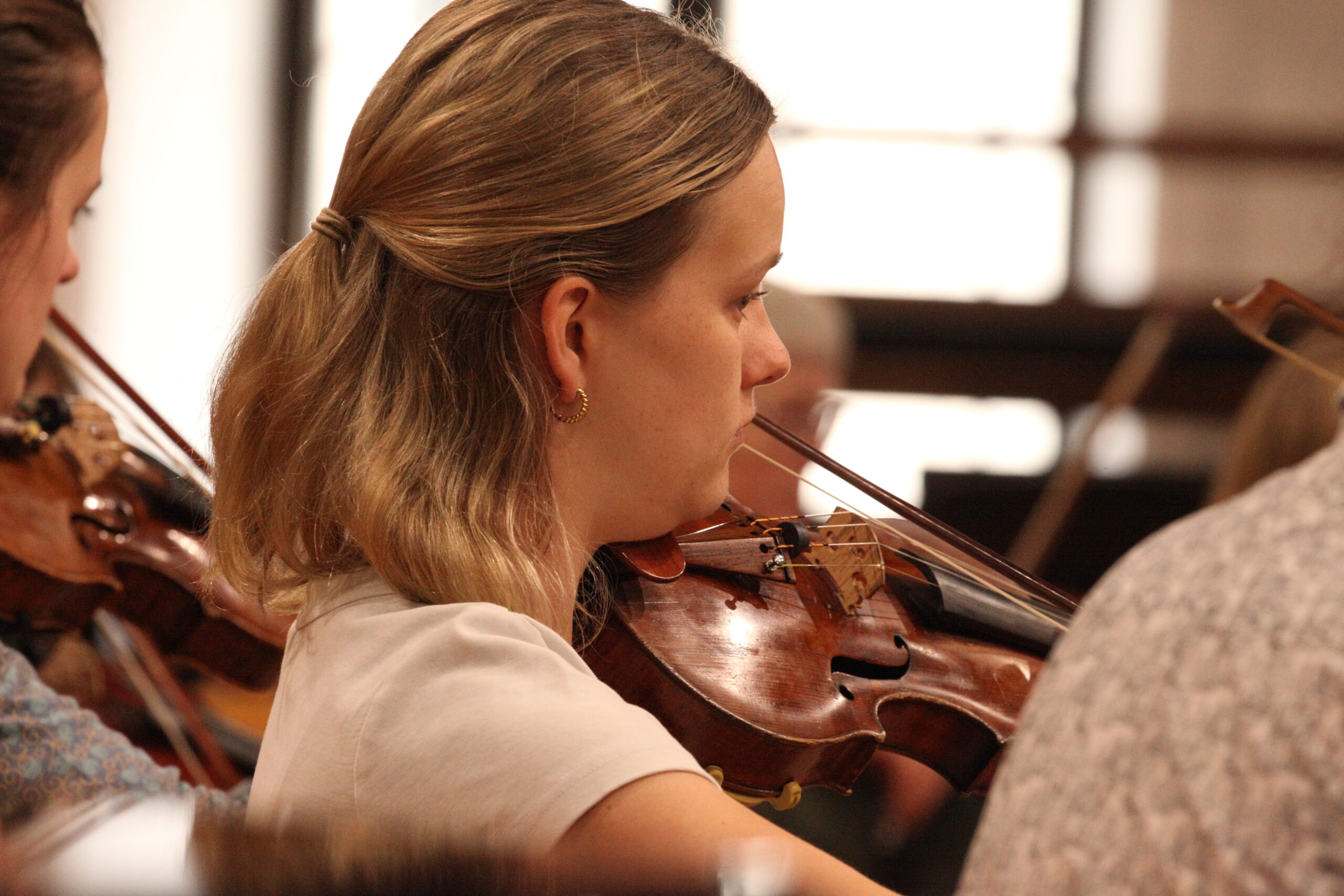 A violin student, pictured in profile, playing in rehearsal, surrounded by other violinists.