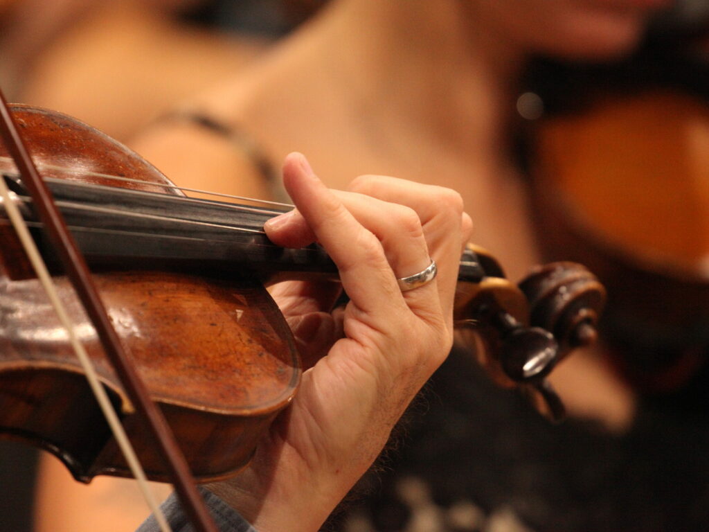 A close-up of a violinist's hand on the fingerboard, fingers pressing down on strings and little finger pointing up.