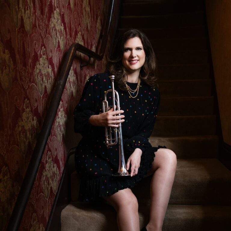 Trumpet player Jo Harris, sitting on a narrow carpeted staircase, next to a wall covered with a lavishly-patterned red-and-gold velvet wall paper and a handrail. She is wearing a short black dress with small multicoloured dots. Her instrument is in her right hand, its bell resting on her thigh. Jo is smiling at the camera. The staircase is very dimly lit apart from the light falling on Jo and the wall paper next to her, creating a dramatic effect.