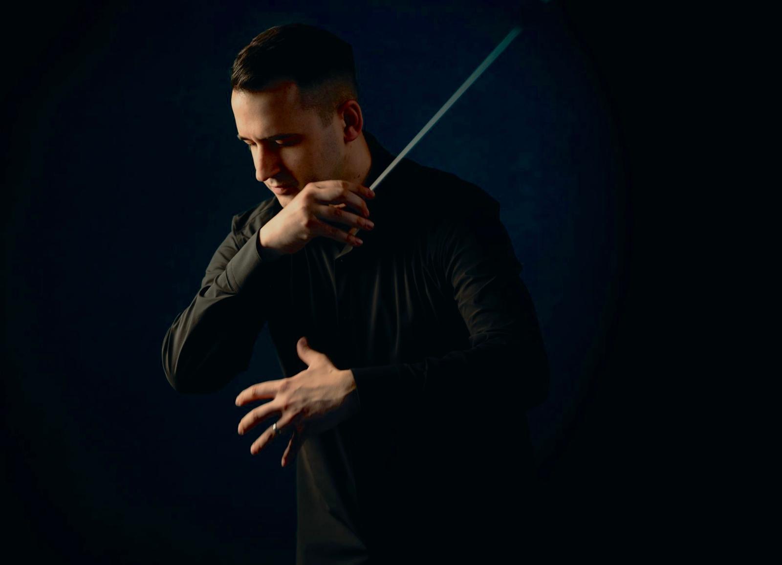 An atmospheric photograph of Johann Stuckenbruck, a dark-haired man, in a black shirt striking a conducting pose against an ink-blue background. His hands and one side of his face are lit by a sideways light. His eyes are closed, his baton hand raised across his body and the fingers of his other hand splayed with expression.