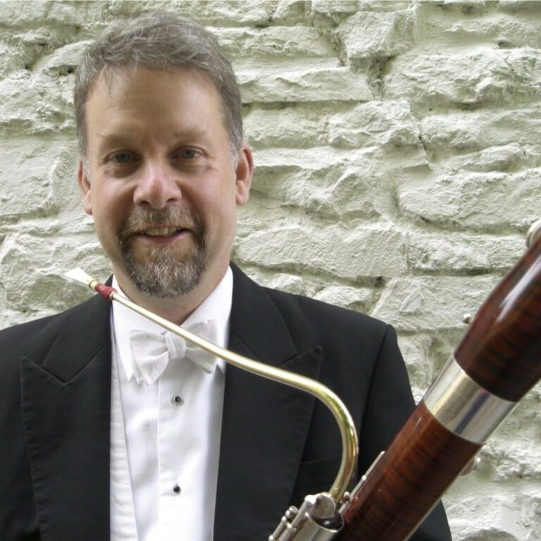 A headshot of Meyrick Alexander, pictured against a whitewashed brick wall, wearing white tie and holding his bassoon.