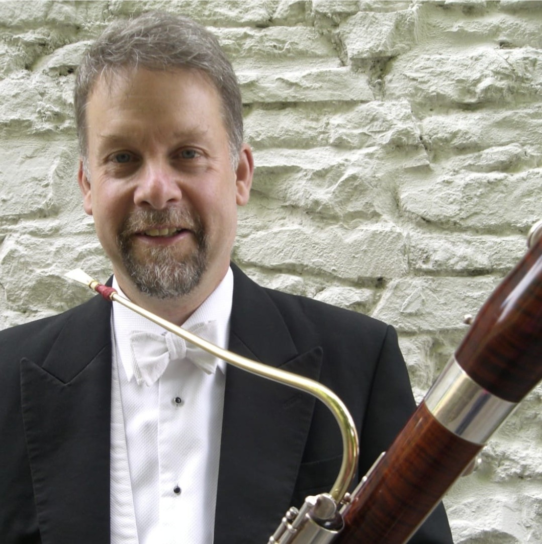A headshot of Meyrick Alexander, pictured against a whitewashed brick wall, wearing white tie and holding his bassoon.