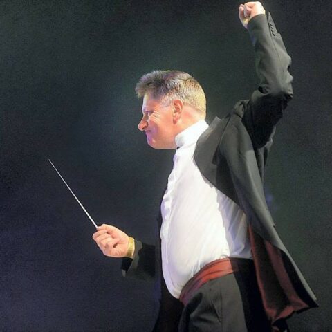 Stephen Bell conducting with aplomb, his baton raised and his left fist punching the air, against a dramatic background of stage smoke and lit by stagelights.