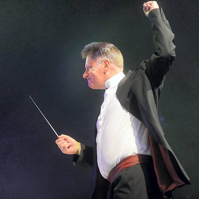 Stephen Bell SQUARE Stephen Bell conducting with aplomb, his baton raised and his left fist punching the air, against a dramatic background of stage smoke and lit by stagelights.