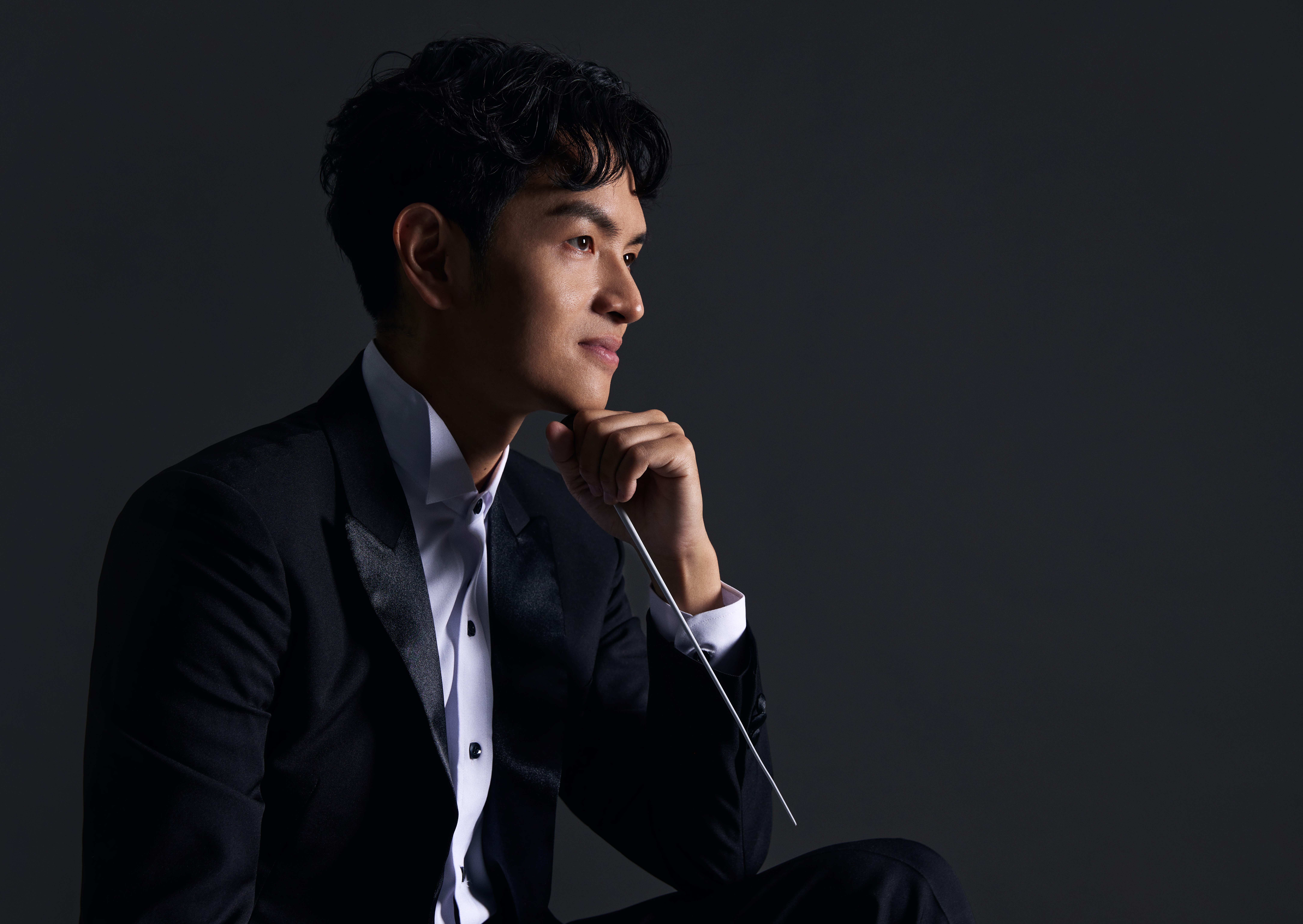A sideways half-portrait of Yi-Chuan Chen. His left elbow rests on his thigh, and his chin on his hand, which holds the baton, downwards, in a casual grip. His face, hand and shirt front are lit by a light source from the right, in dramatic contrast to the shadow created elsewhere and dark grey background.