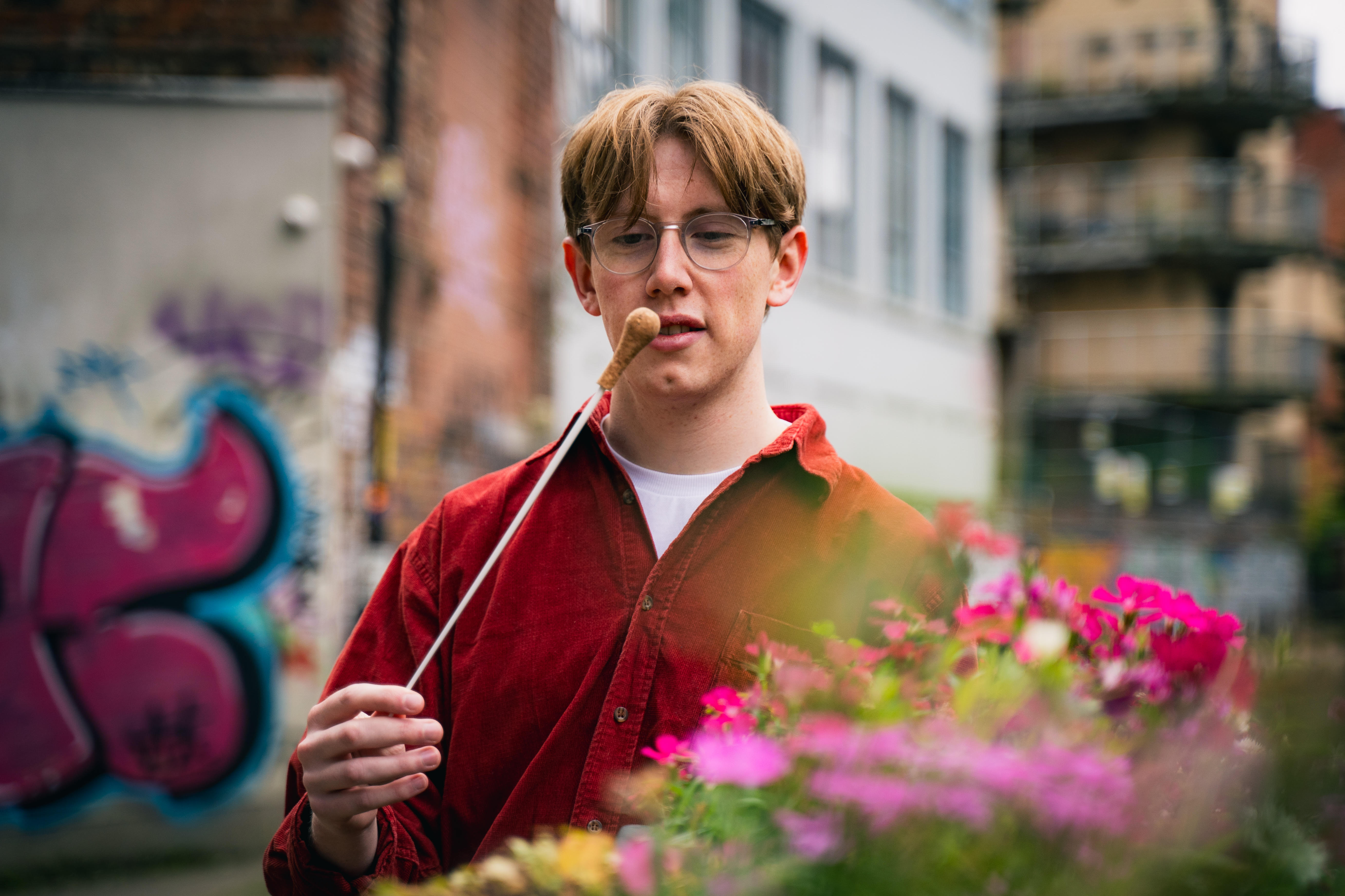 A half-portrait of Benjamin Draper in an urban street, holding his baton upside down by the tip, seemingly contemplating it. He is wearing a loose bright red shirt, top button open, with a white T-shirt underneath. There are some out-of-focus flowers in a planter in the foreground, and some graffiti on a wall in the background.
