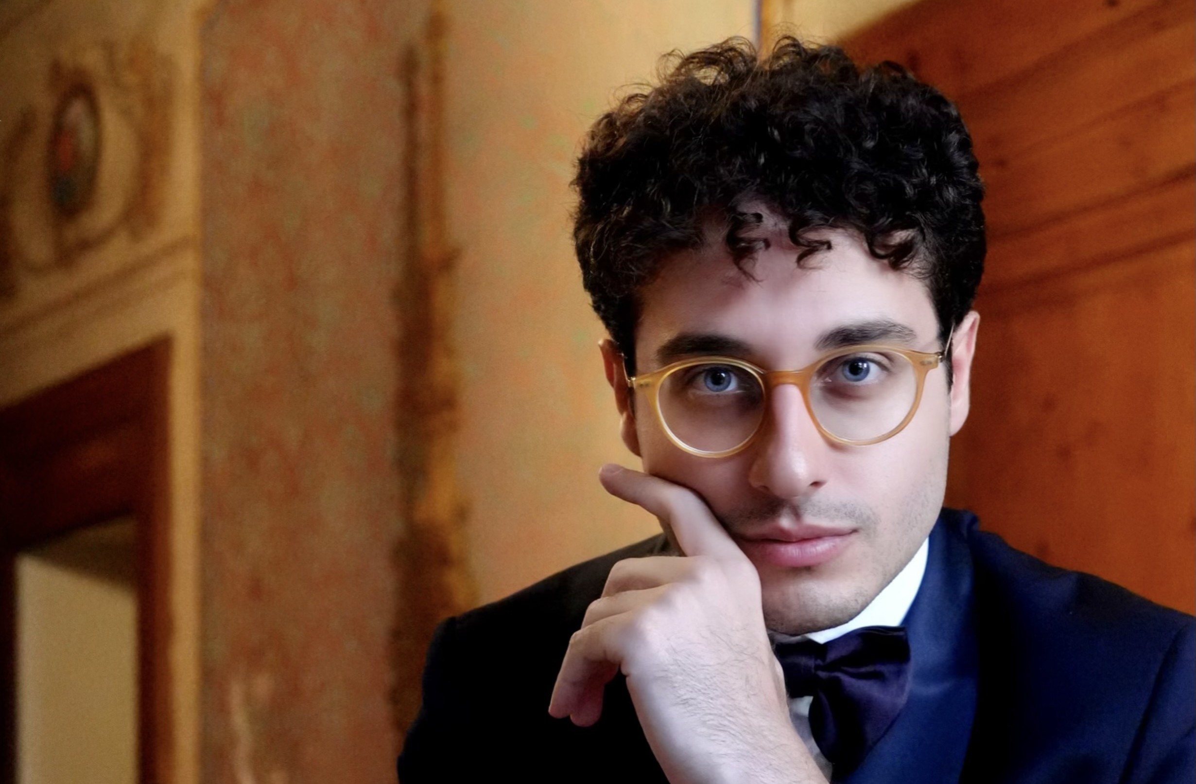 A photo portrait of conductor Giuseppe Stillitano in an eighteenth-century Italian interior. His left hand is raised, index finger across his right cheek. He is is gazing directly into the camera. He is wearing an ink blue suit with a crisp white shirt and a bow tie. The frame of his glasses picks up the honey shades of the background.