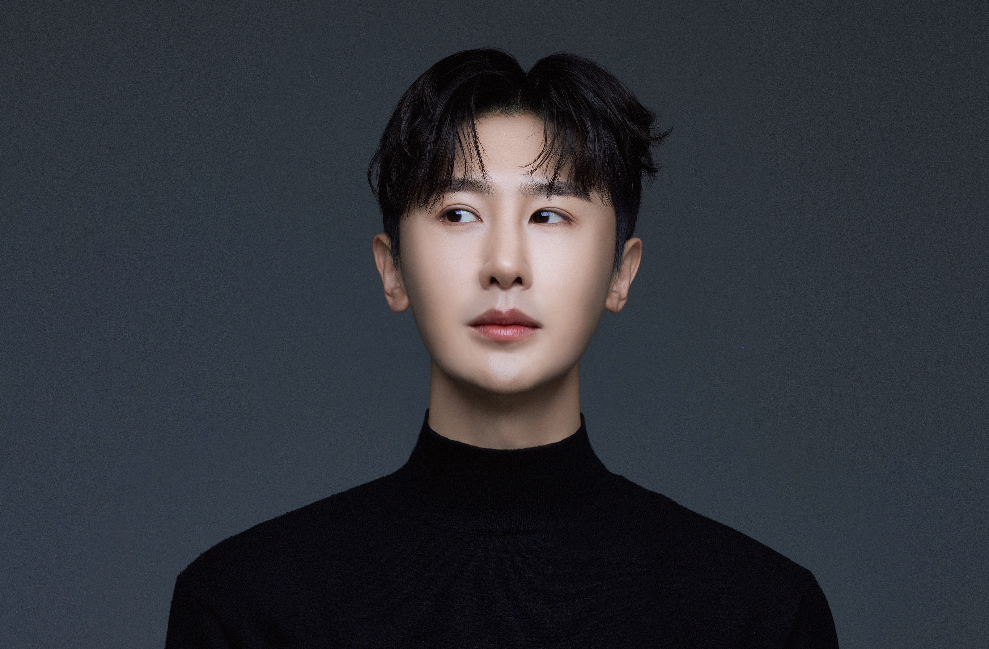Promotional headshot of Korean conductor Wooju Baik, dressed in all-black turtleneck against a grey background. He is facing the camera directly but looking to his right with a very sideways glance.