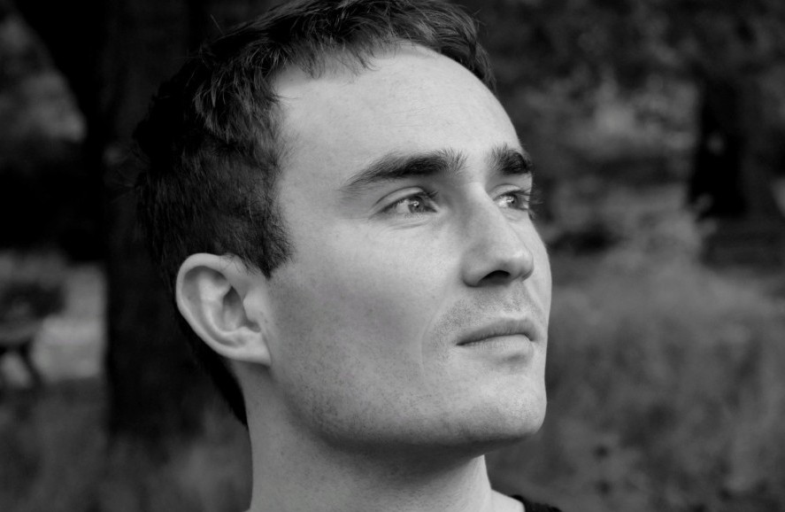 A black-and-white headshot of Michael Coleby. He is gazing into the distance to his left, wearing a black T-shirt, against a parkland background.