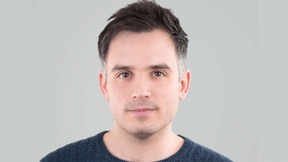 Headshot of conductor Robert Hodge. He is gazing into the distance behind the camera, wearing a dark blue cable-knit jumper, against a blank pale grey background.