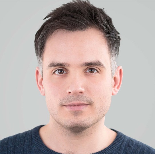 Headshot of conductor Robert Hodge. He is gazing into the distance behind the camera, wearing a dark blue cable-knit jumper, against a blank pale grey background.