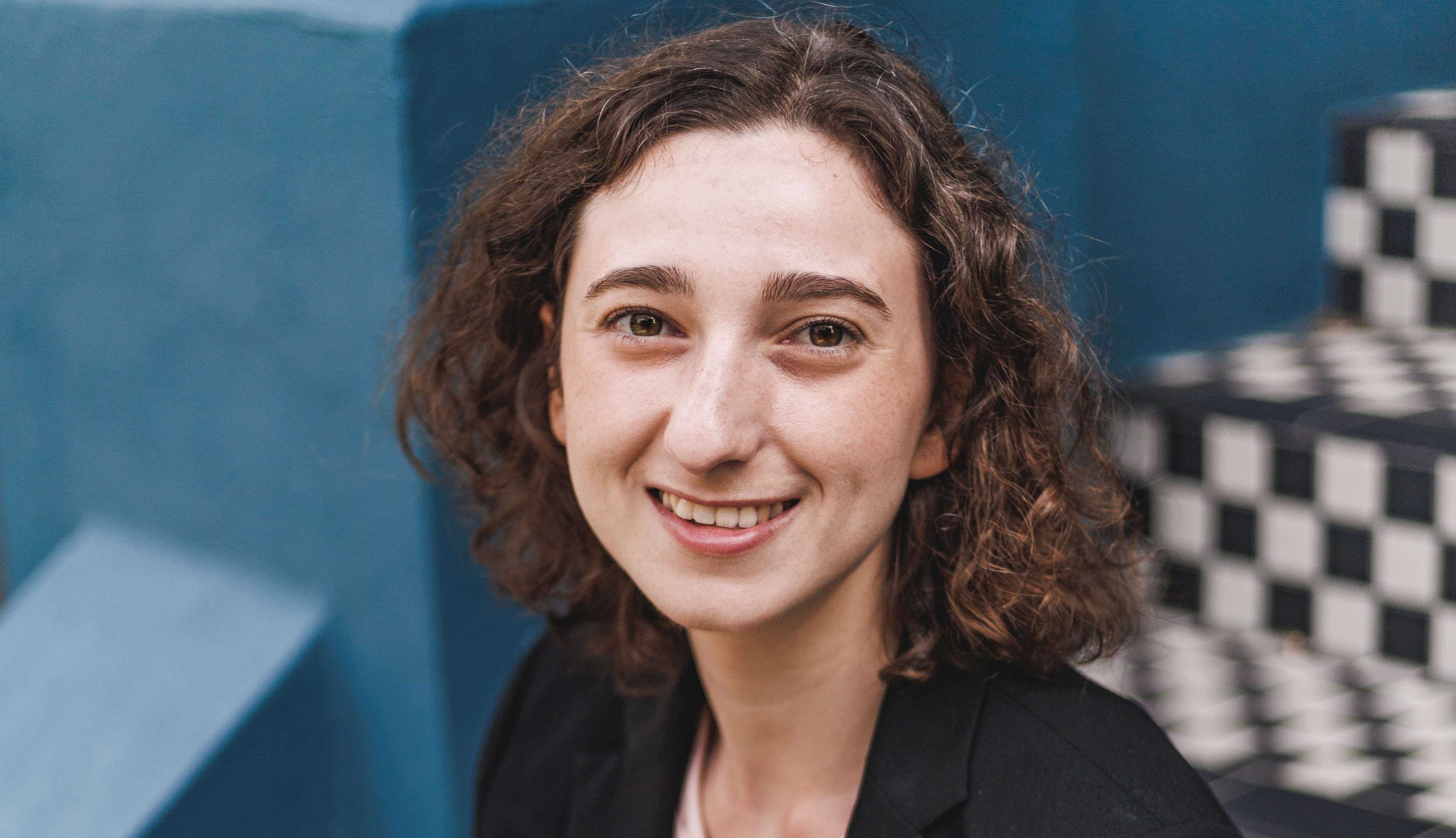 A headshot of Michal Oren who is smiling at the camera with an open gaze, her face framed by her curly brown hair. She is sitting, at a slight angle to the camera, on some black-and-white chequerboard steps, visible in the background, with a teal-coloured wall behind her.