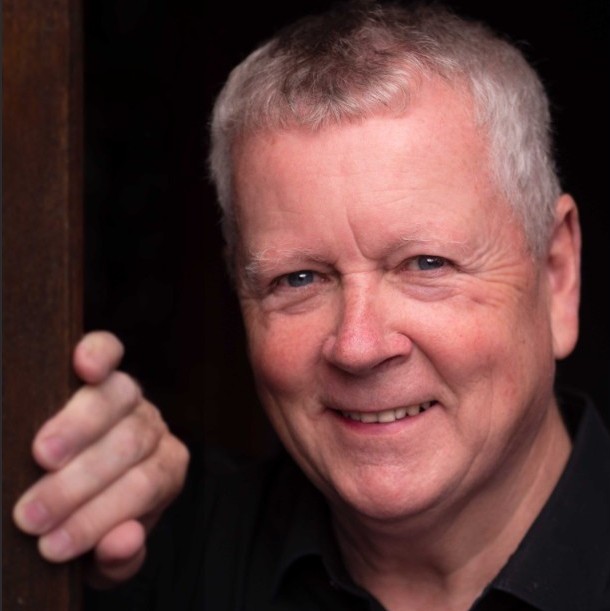 Headshot of Roger Coull, who is looking into the camera with a twinkly smile. He is wearing a black shirt against a dark background, with his right hand resting on a door frame at chin height.