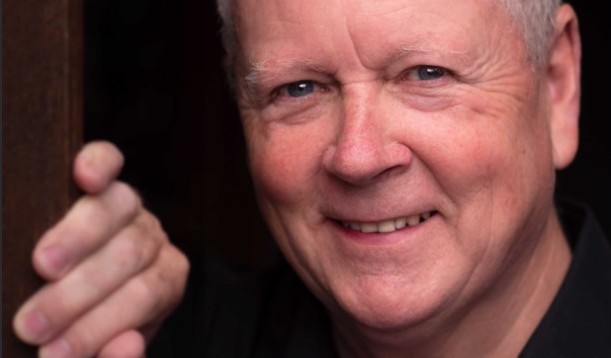 A cropped headshot of Roger Coull, who looking into the camera with a twinkly smile. He is wearing a black shirt against a dark background, with his right hand resting on a door frame at chin height.