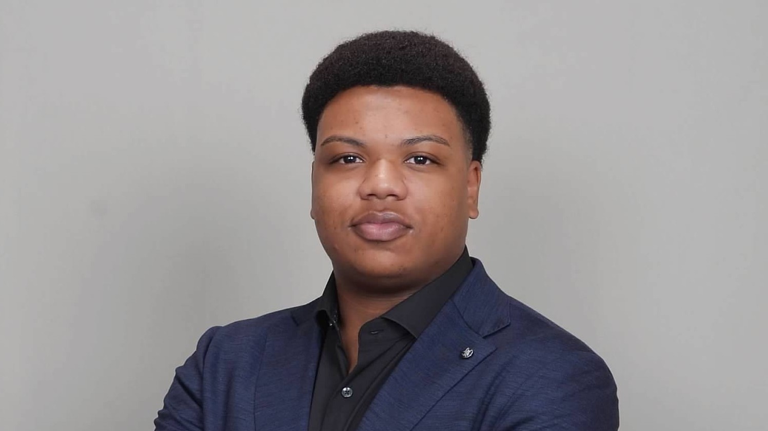 A headshot of Maliq Veal, who is looking directly into the camera with an air of steady self-assurance. His shoulders are slightly angled and he is wearing a black shirt with the top button open under an indigo blue jacket with a lapel pin.