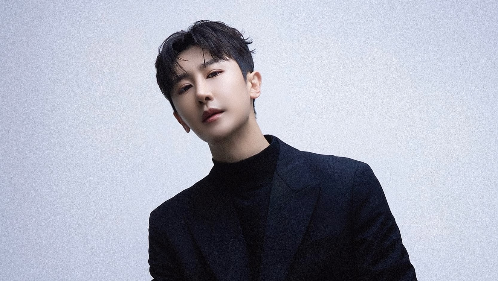 A headshot of Wooju Baik, who is leaning over to his right and looking into the camera with a slightly aloof air. He is wearing a black turtleneck jumper and a very dark navy jacket.