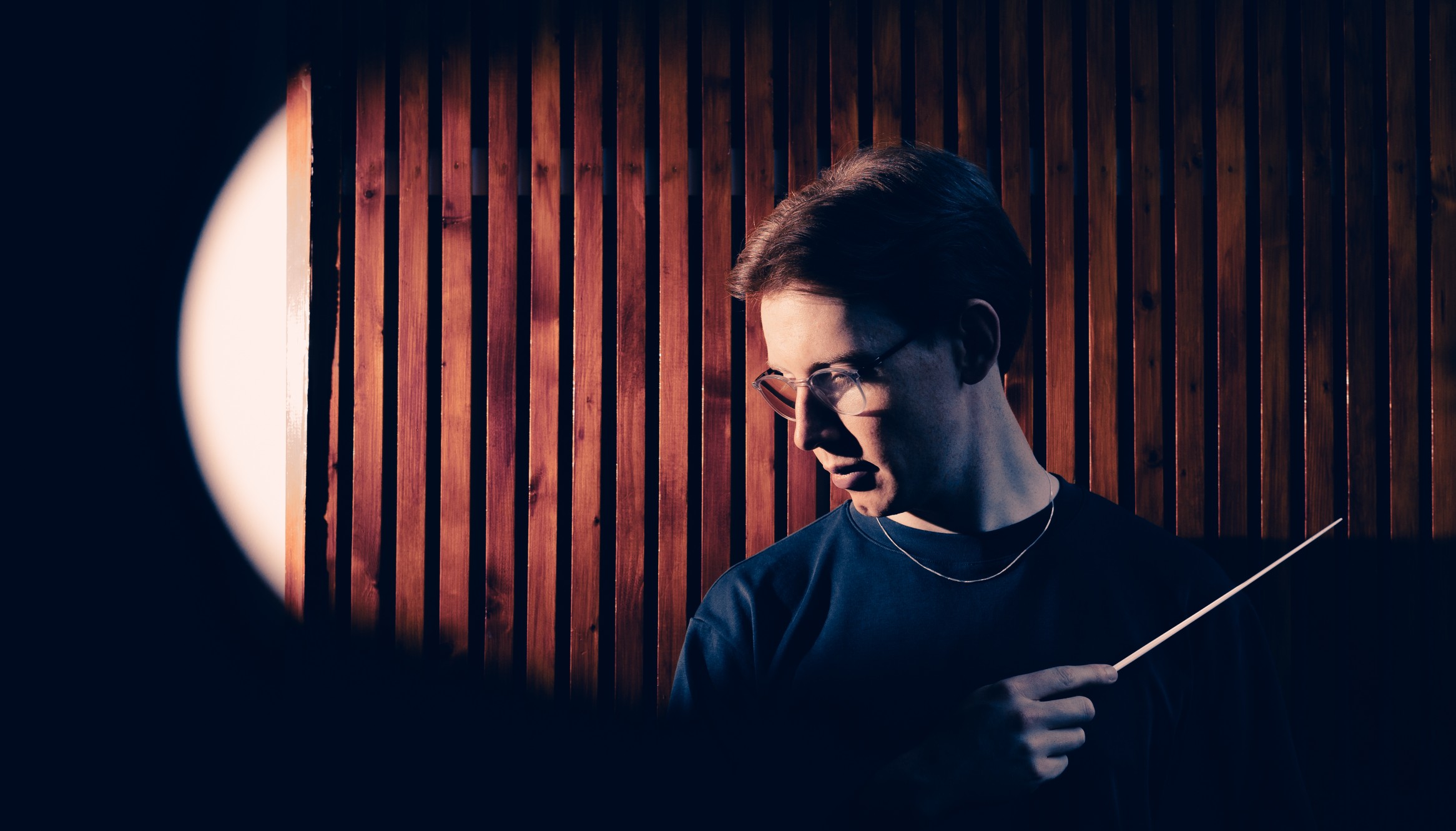 A headshot of Benjamin Draper, who is leaning against a wall panelled with dark honey-coloured wooden slats, gazing down to his right deep in thought. He is holding his baton, slightly raised, in his right hand which is resting against his chest. He is wearing a dark O-neck jumper and a thin chain. The camera captures him in a sideways light which casts shadows across the side of his face, viewed through an oval opening in a door (or similar), creating a spotlight effect.