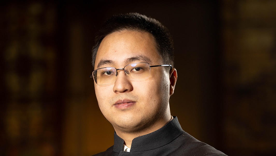 A headshot of Zihan Zhou, who is gazing steadily into the camera through his half-framed glasses. He is wearing a black formal jacket with a mandarin collar, a white shirt just visible underneath. The light is warm and the background has rich brown tones into which the outline of his short hair partly disappears.