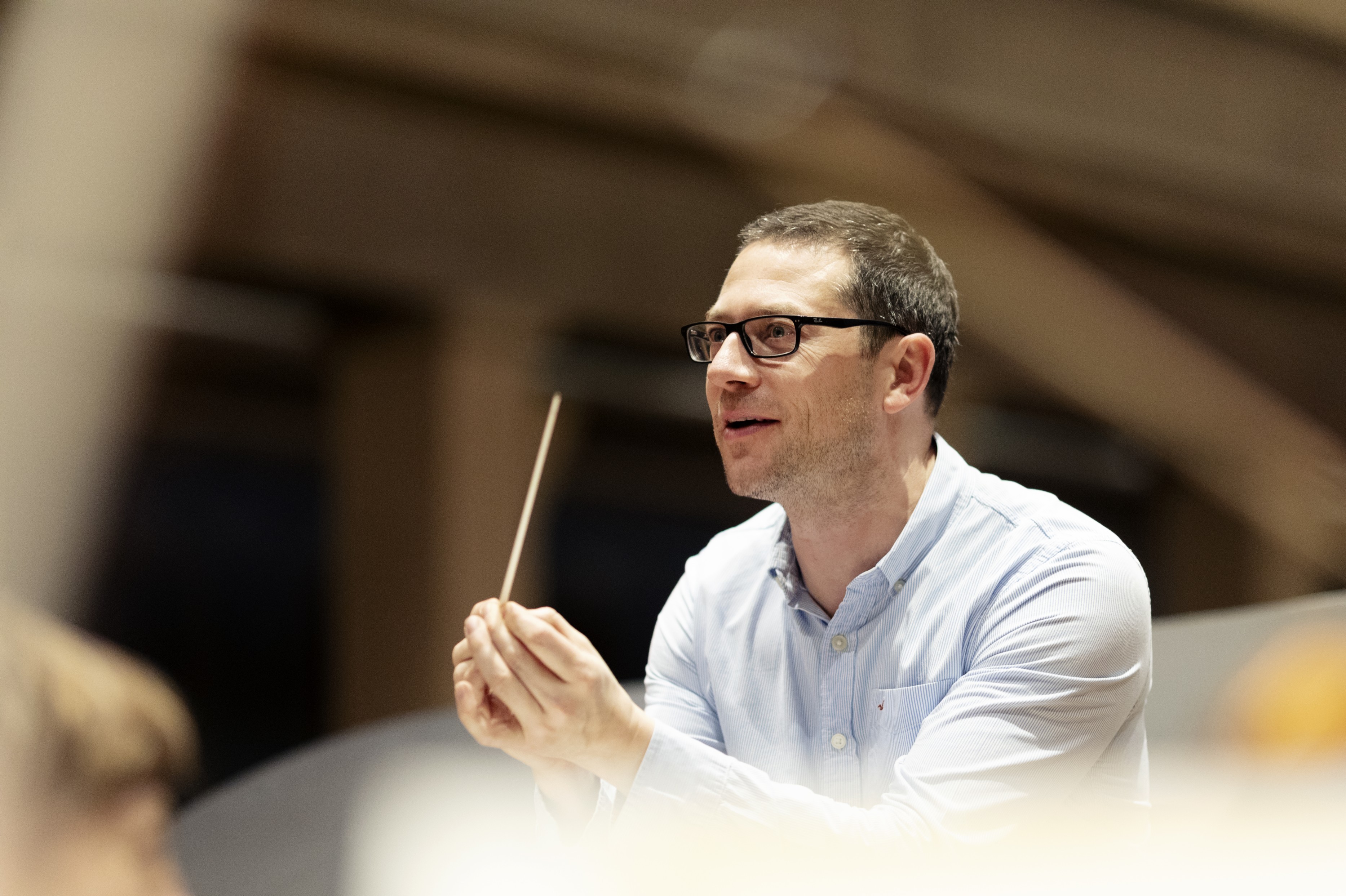 Conductor John Wilson at rehearsal, holding his baton raised in both hands, ready to conduct, with a focused and excited expression. He is raring to go with the music.
