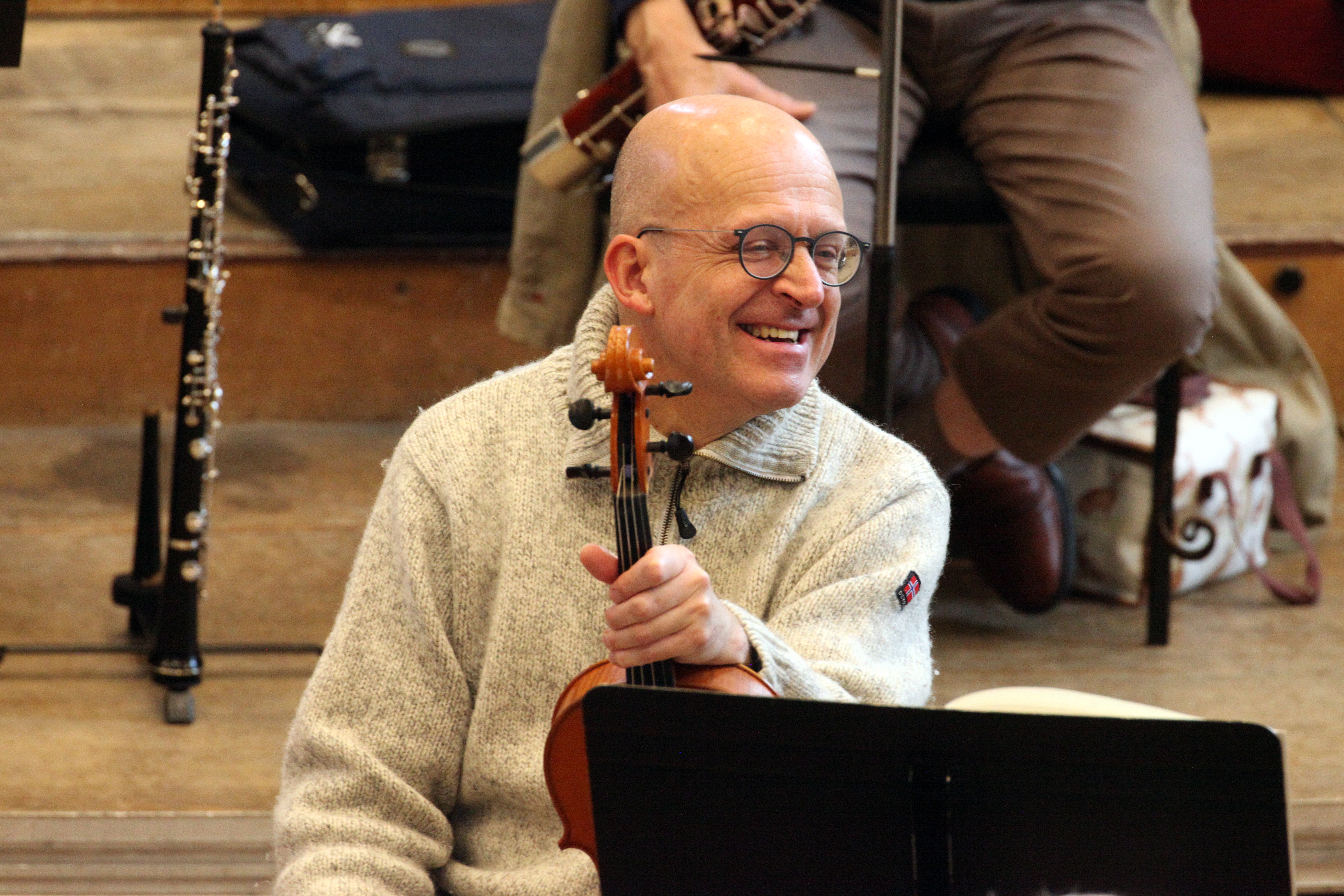 A viola player during a break in play at rehearsal, laughing. They are resting their instrument on their knee, holding it by the neck close to the body of the instrument.