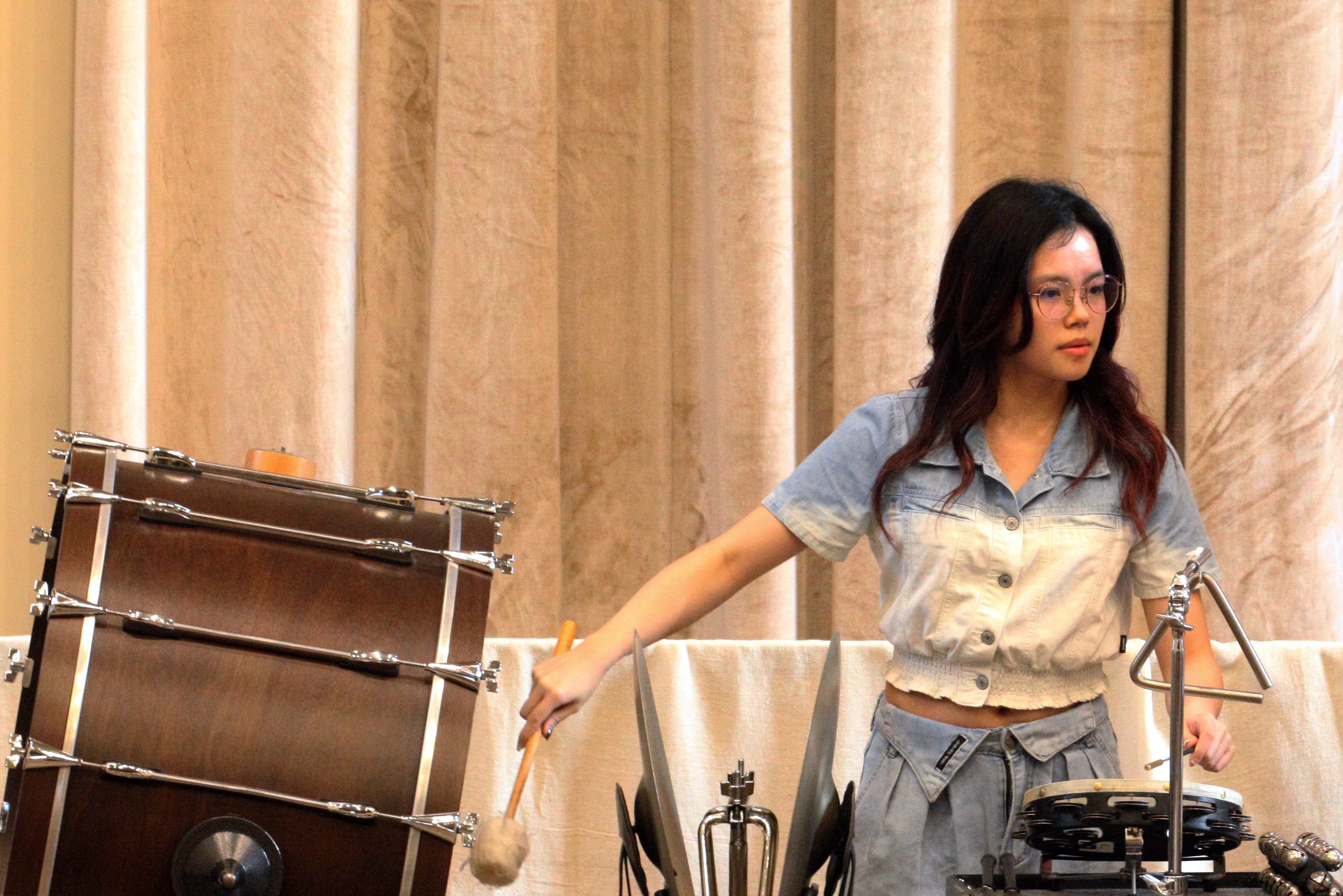 A student percussionist playing bass drum, triangle and tambourine concurrently, with an intent gaze on the part, with cymbals and sleigh bells also in front of them.