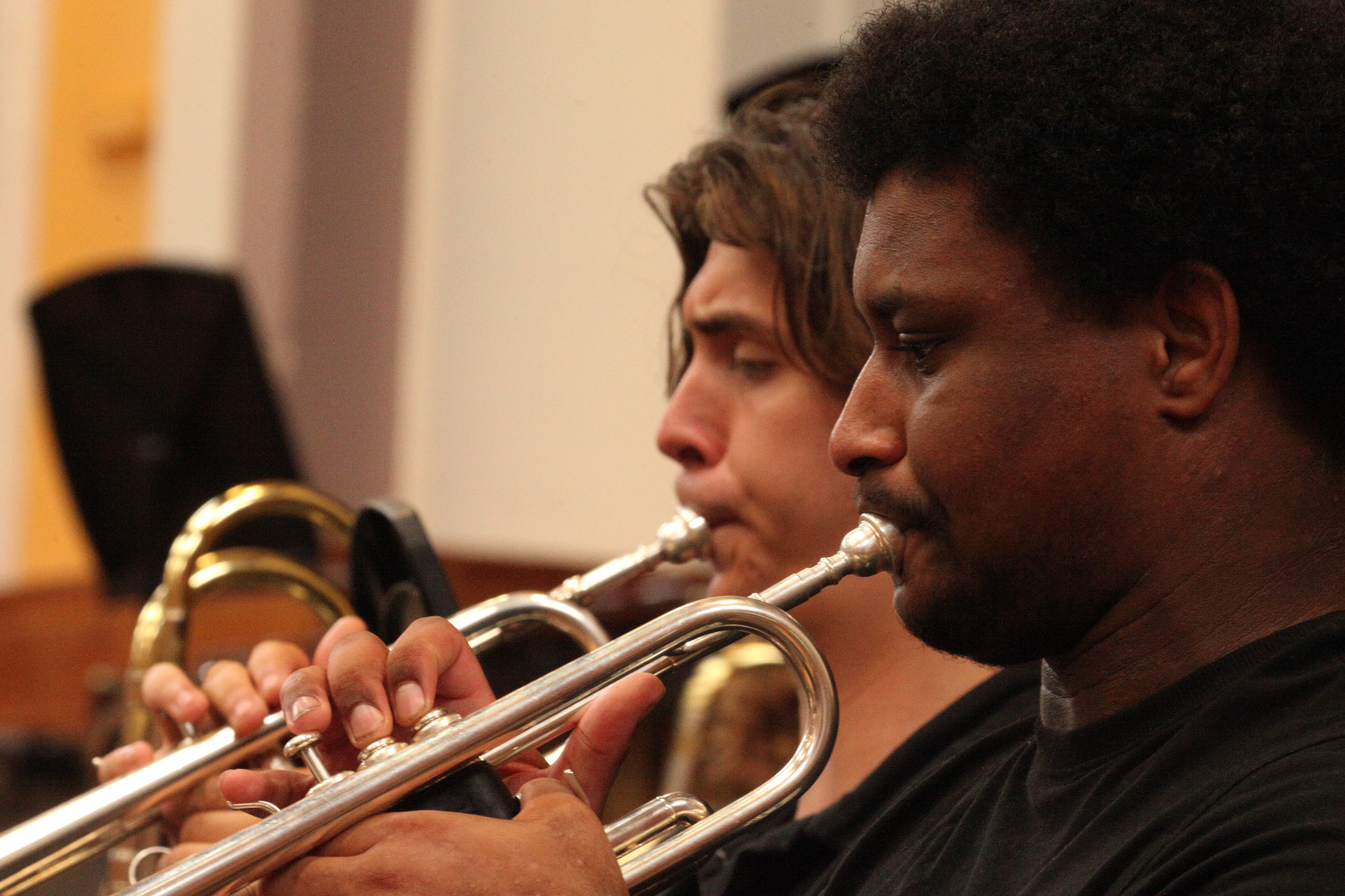Two student trumpet players in rehearsal, with low brass instuments in the background. Lights make the brass of the instruments shine.